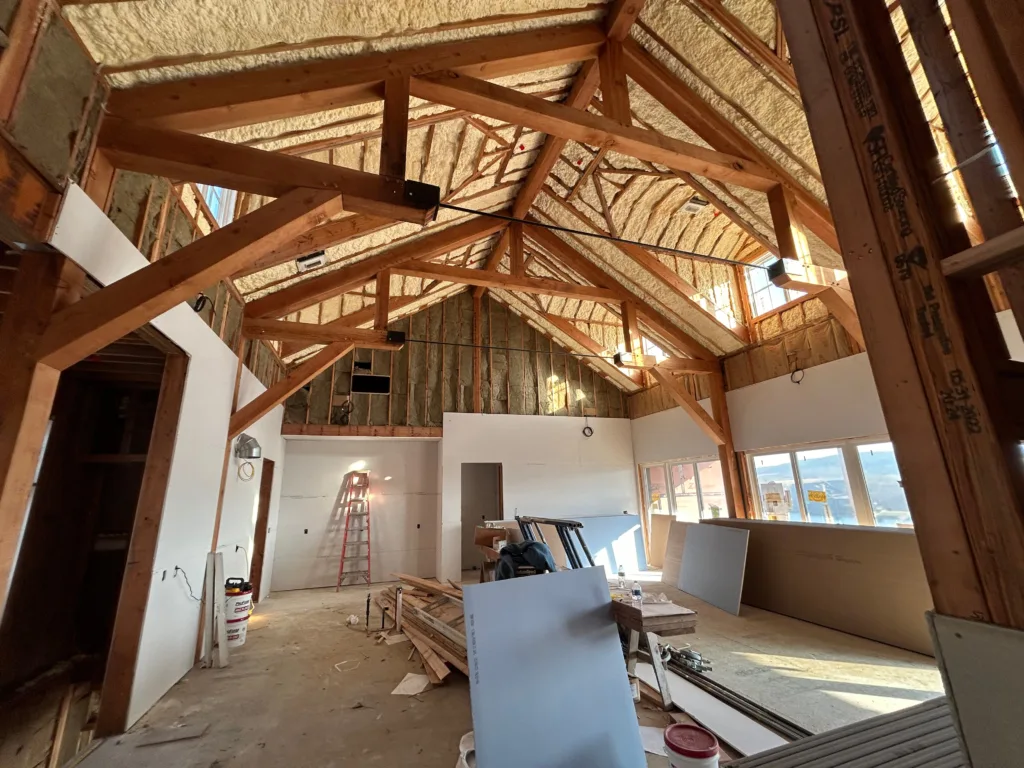 Construction site view of a custom home drywall installation, showing stacked gypsum boards on the floor and exposed timber ceiling trusses.