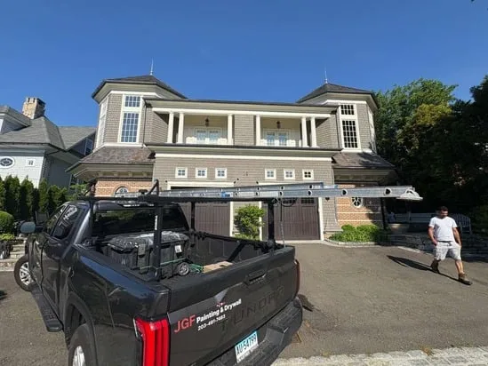 Contractor truck equipped with ladders parked in the driveway of a large home during an exterior house painting project.