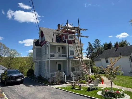 Wide street view of a historic home undergoing Victorian exterior turret painting, displaying the full scaffolding structure built around the front tower.